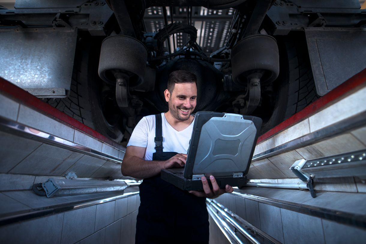 Technician running diagnostics on a rugged laptop beneath a heavy vehicle